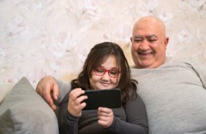 Bestefar og barnebarn sitter i sofa og flirer mens de ser på noe på mobilen